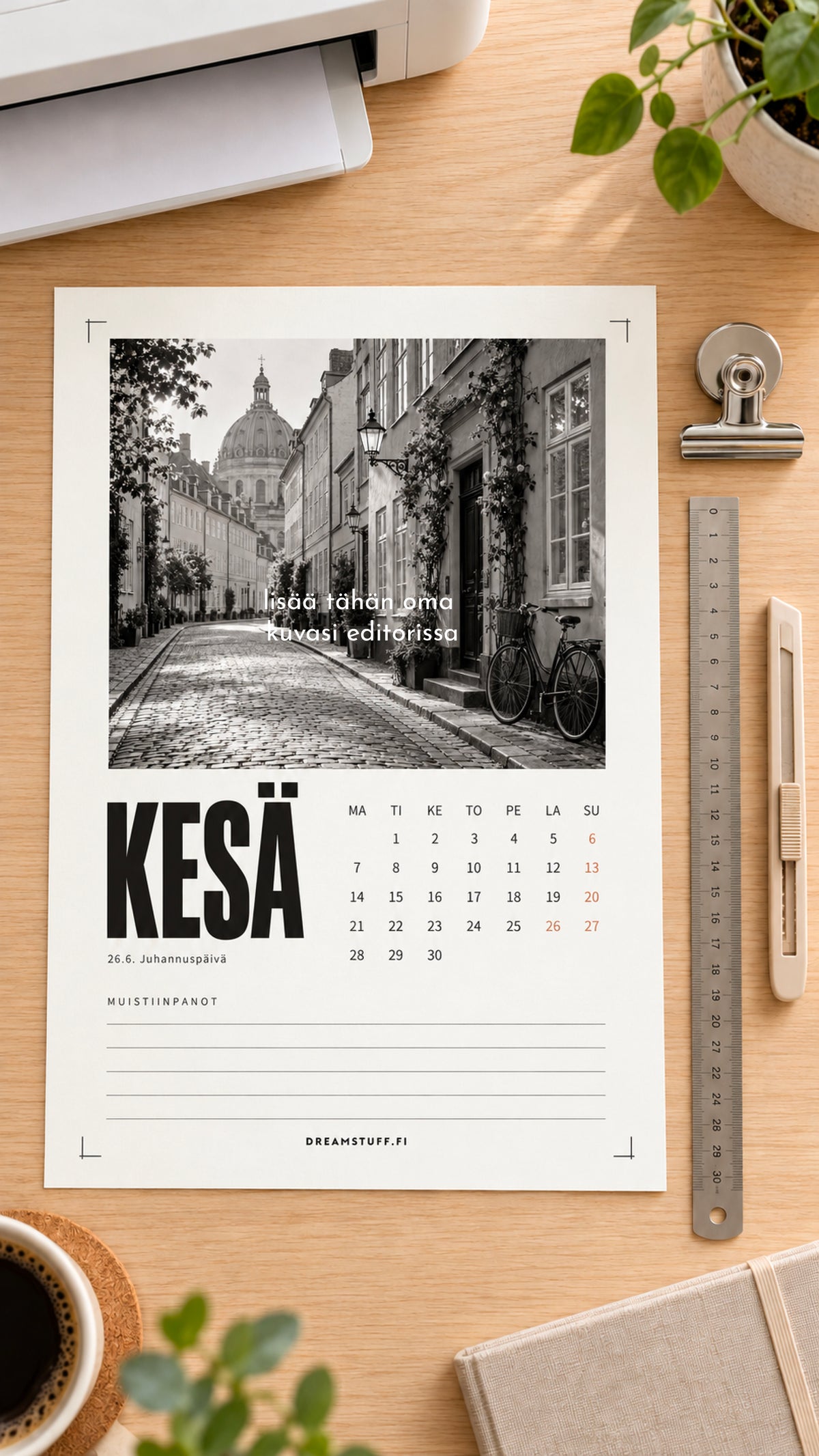 Tulostettava valokuvakalenteri 2027 A4-arkilla leikkuumerkkeineen kotitulostimen vieressä, kesäkuun sivulla mustavalkoinen kaupunkikuva.
