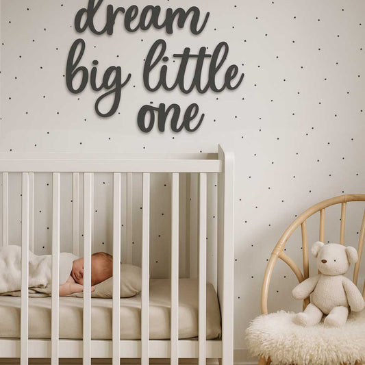 Koivuvanerinen musta "dream big little one" -tekstikoriste lastenhuoneen seinällä, valkoinen sänky ja korituoli skandinaavisessa sisustuksessa.