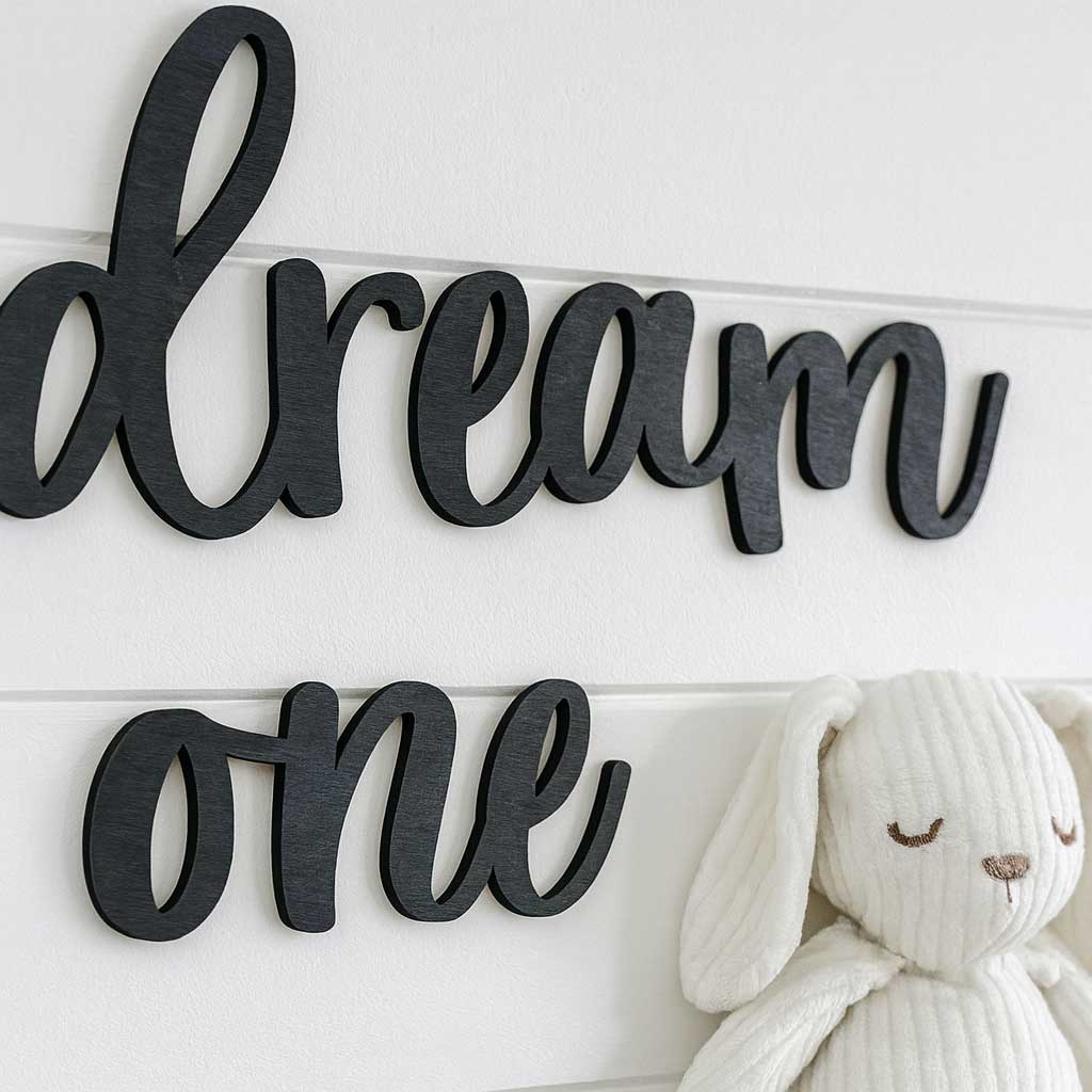 Mustasta koivuvanerista laserleikattu teksti "dream" ja "one" valkoisella lautaseinällä, vieressä valkoinen skandinaavinen pupupehmolelu