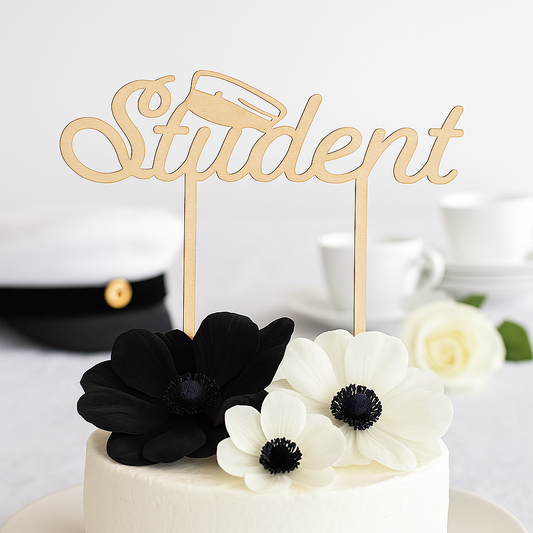 Dreamstuff vaalea koivuvanerinen Student-kakkukoriste valkoisen juhlakakun päällä, koristeena mustia ja valkoisia kukkia. Taustalla ylioppilaslakki, valkoinen ruusu ja kahvikuppi.