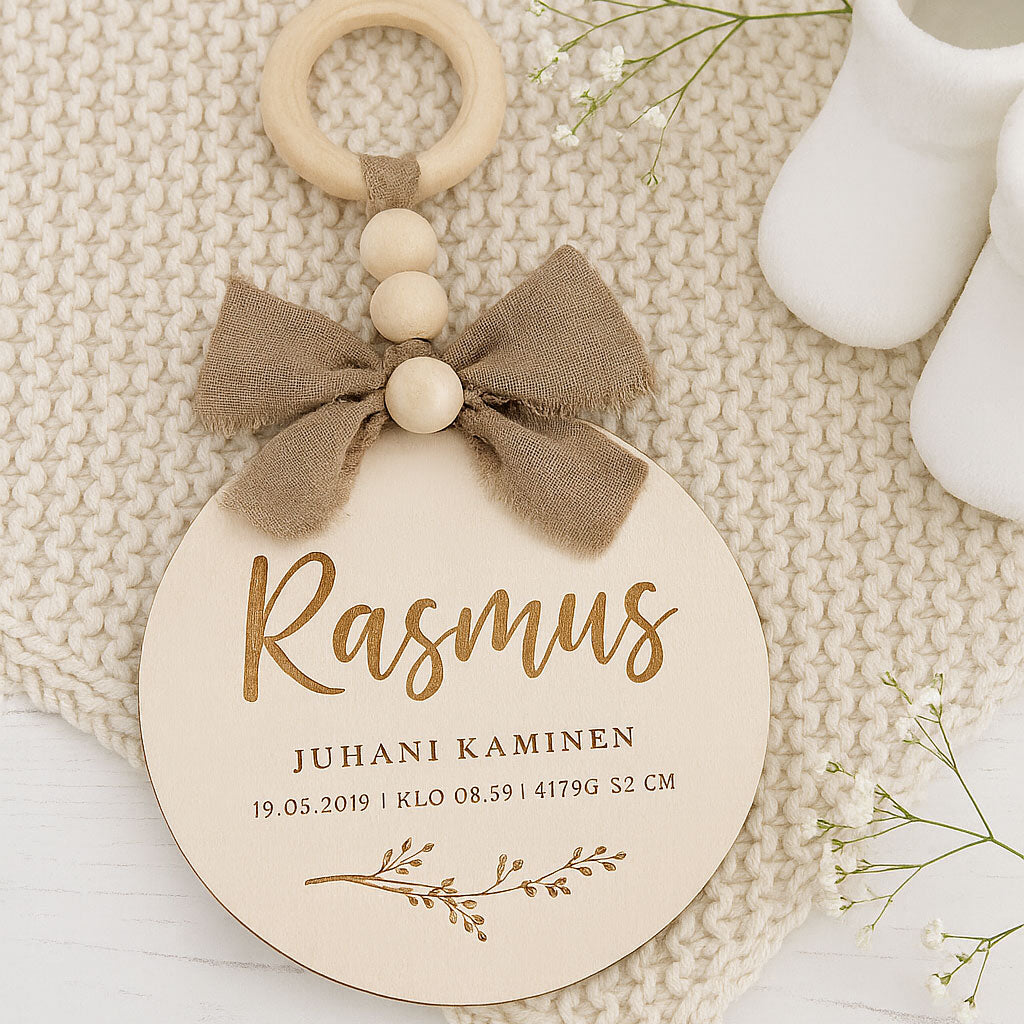 Dreamstuffin personoitu nimikyltti vauvalle – koivuvanerinen kyltti beigerusetilla ja puuhelmillä, asetettuna pehmeälle neulealustalle vauvanvaatteiden viereen.