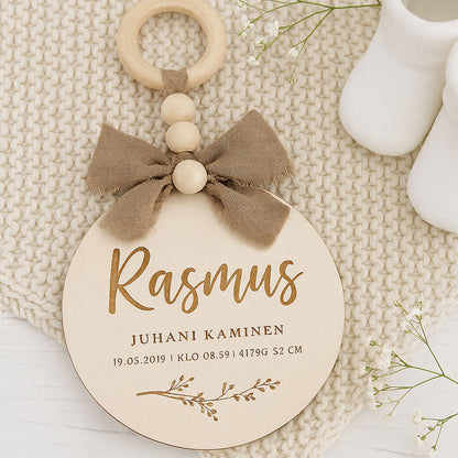 Dreamstuffin personoitu nimikyltti vauvalle – koivuvanerinen kyltti beigerusetilla ja puuhelmillä, asetettuna pehmeälle neulealustalle vauvanvaatteiden viereen.
