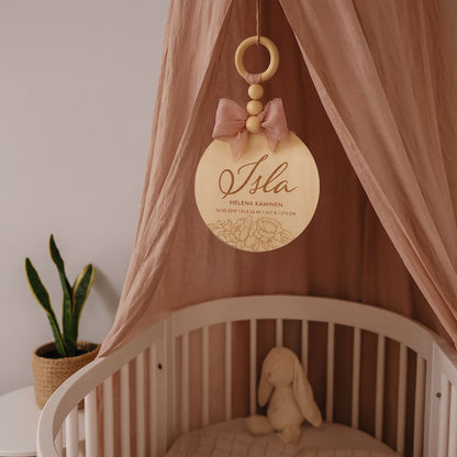 Dreamstuffin personoitu nimikyltti vauvalle – koivuvanerinen kyltti vaaleanpunaisella rusetilla ja puuhelmillä, ripustettuna pinnasängyn yläpuolelle lastenhuoneen sisustuksessa.