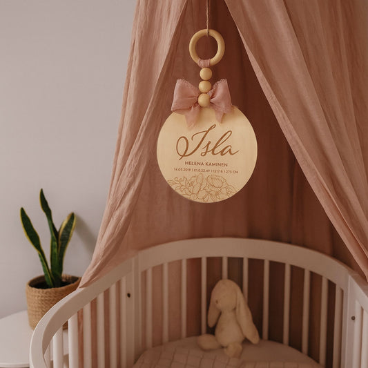 Dreamstuffin personoitu nimikyltti vauvalle – koivuvanerinen kyltti vaaleanpunaisella rusetilla ja puuhelmillä, ripustettuna pinnasängyn yläpuolelle lastenhuoneen sisustuksessa.